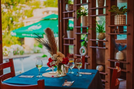 Masă romantic aranjată cu un pahar de vin și decorațiuni florale lângă fereastră la restaurantul Dukov House, orașul Kavarna